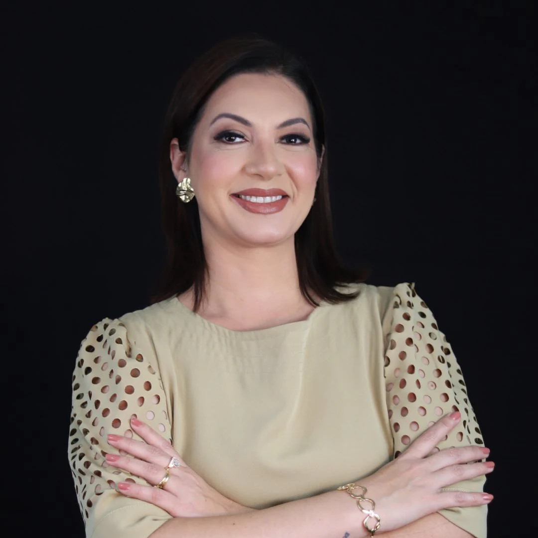 Dra. Cíntia Urquiza, fisioterapeuta dermatofuncional, de braços cruzados, sobre fundo escuro
